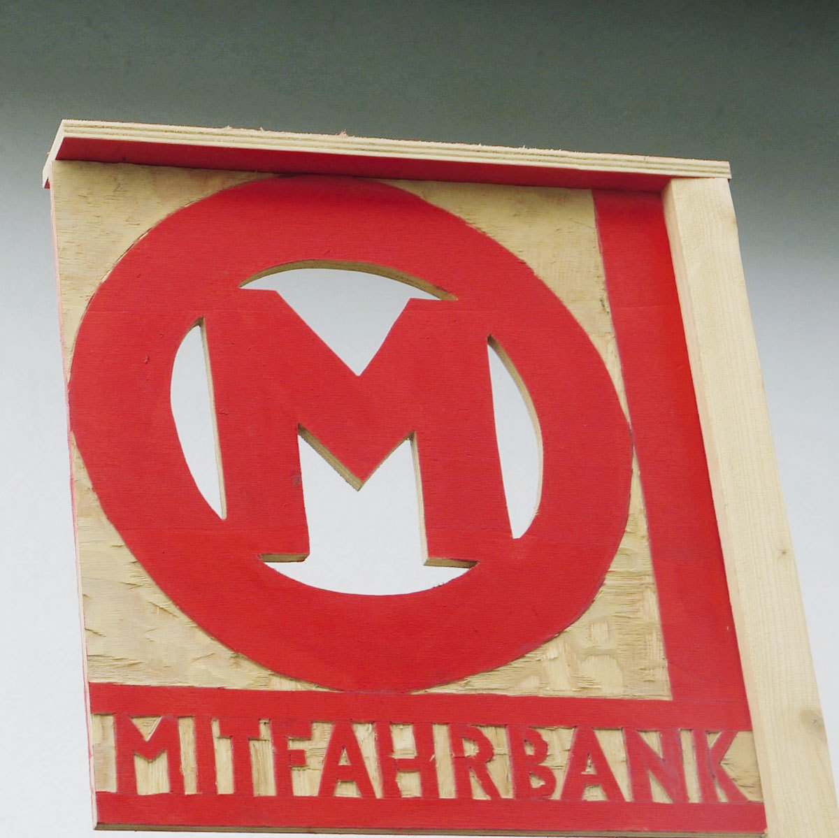 Eine rechteckige helle Holzplatte ist mit ausgesägtem Logo und Buchstaben versehen. Oben prangt in Rot der Buchstabe »M« in einem roten Kreis mit weißer Füllung, unten ist zwischen zwei roten Linien das Wort »MITFAHRBANK« zu lesen.