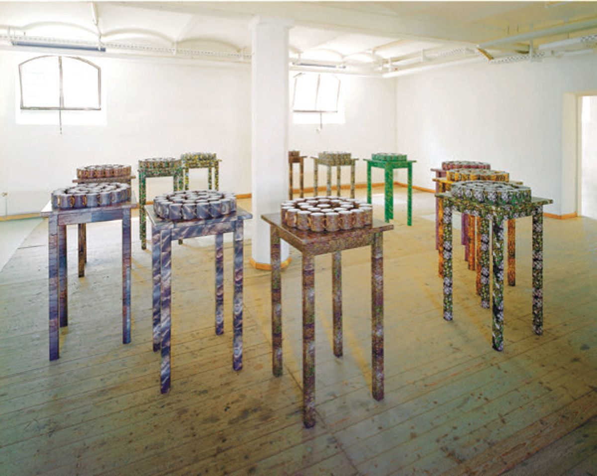 In einem Ausstellungsraum stehen kreisförmig zwölf Skulpturen um eine weiße Säule. Die unterschiedlich farbigen Objekte haben die Form von hohen Sitzstühlen ohne Lehne, auf der Sitzfläche ist jeweils ein tortenähnlicher Kreis angebracht.