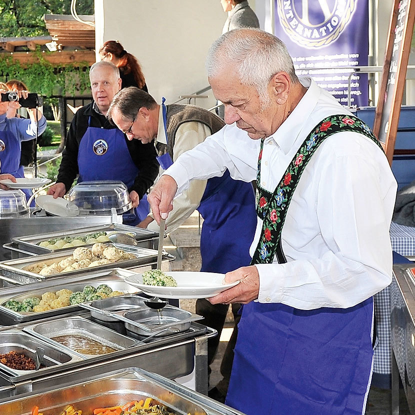 Mehrere Personen mit blauen Schürzen stehen vor Edelstahl-Speisenwärmern und verteilen Essen auf Tellern.