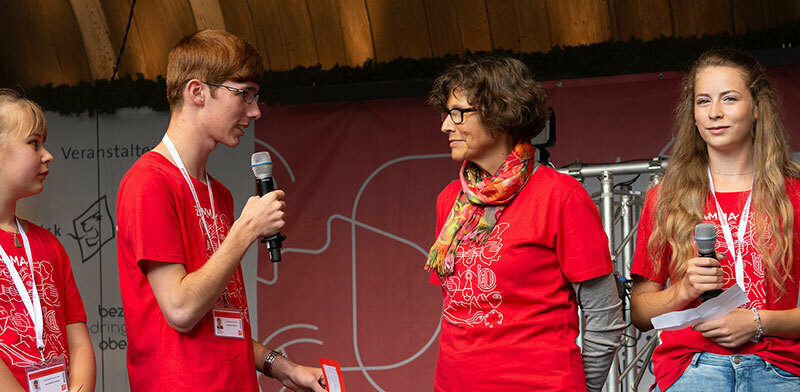 Ein junger Mann interviewt eine Frau auf einer Bühne. Links und rechts daneben steht jeweils eine junge Frau. Alle tragen rote T-Shirts mit dem Aufdruck "Zamma".
