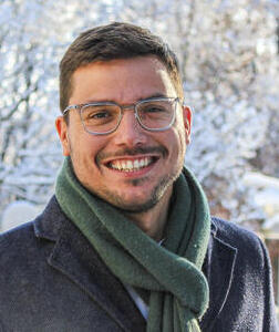 Foto eines jungen, freundlich lachenden Mannes mit Brille. Im Hintergrund ist eine Schneelandschaft zu erkennen.